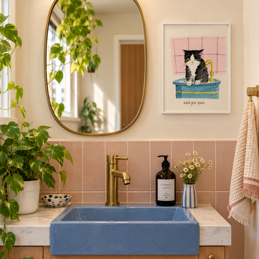 Quadro A4 com ilustração de gato preto e branco e frase “wash your paws”, em moldura branca, ao lado do espelho em lavabo com azulejos rosa e pia azul.